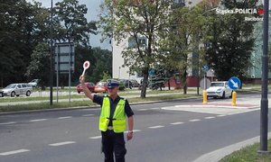 zdjęcie przedstawia policjanta z uniesioną ręką. Policjant ubrany jest w białą czapkę i odblaskową kamizelkę.