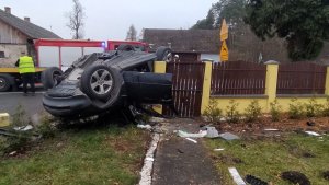 Na zdjęciu widać ciemny  samochód położony na dachu na ogrodzeniu betonowym. Na drugim planie stoi samochód straży pożarnej oraz laweta.