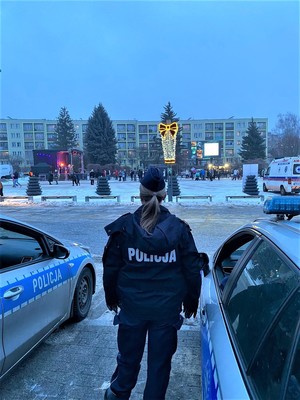 Na zdjęciu Policjantka na tle Placu Tysiąclecia w Chrzanowie, stoi pomiędzy radiowozami. Na dalszym planie stoiska związane z 34 Finałem WOŚP.