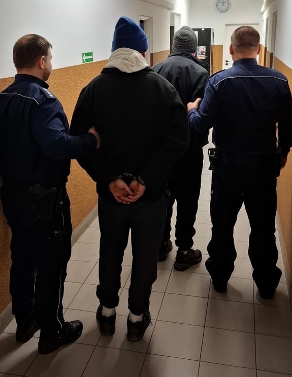 Policjanci stoją z zatrzymanymi mężczyznami. Mężczyźni mają nałożone kajdanki na ręce trzymane z tyłu.