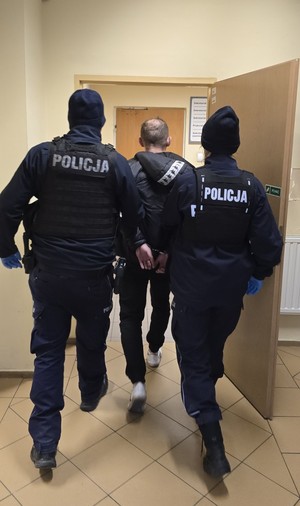 Policjanci Ogniwa Parolowo-Interwencyjnego KPP Chrzanów prowadzą zatrzymanego 38-latka do Prokuratury Rejonowej w Chrzanowie. Zatrzymany ma założone kajdanki na ręce trzymane z tyłu.