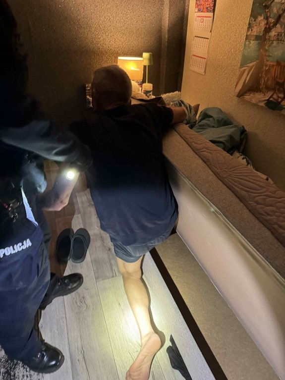 Mężczyzna wychodzący z wersalki, obok stoi policjant, który w ręku trzyma latarkę i pomaga mężczyźnie wstać.