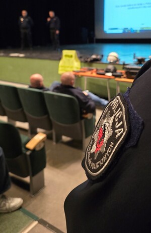 Na zdjęciu widoczne ramię policjanta z naszywką Instruktora Strzelań Policyjnych. W rozmazanym tle, osoby siedzące na widowni oraz referujące.