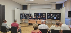 Uczestnicy spotkania siedzący na widowni oraz policjanci siedzący za stołem na podwyższeniu.