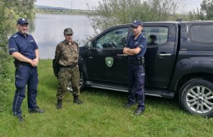 Na zdjęciu dwaj umundurowani policjanci wraz z umundurowanym funkcjonariuszem Straży Rybackiej na tle samochodu Straży Rybackiej.