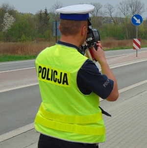 Na zdjęciu widać policjanta wydziału ruchu drogowego ubranego w żółtą kamizelkę odblaskową i białą czapkę wykonującego pomiar prędkości.